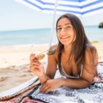 Woman on beach with sunglasses, lying on blanket under umbrella, showcasing summer skincare routine.